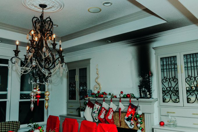 A festive dining room decorated for Christmas, featuring a black chandelier, lit candles, Christmas stockings hung by a fireplace, and red-covered chairs around a table.