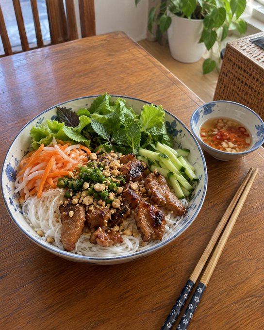 A bowl of Vietnamese vermicelli noodles (bún) topped with grilled pork, fresh herbs, cucumber, and pickled carrots, served with a side of nuoc cham dipping sauce and chopsticks on a wooden table.