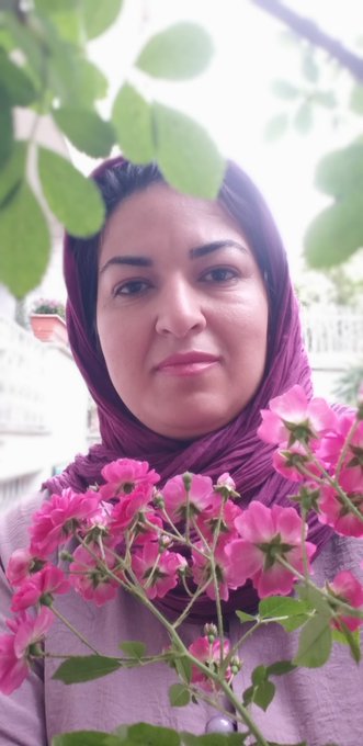 A woman wearing a dark pink headscarf, posing for a portrait with bright pink flowers in the foreground and green leaves above.