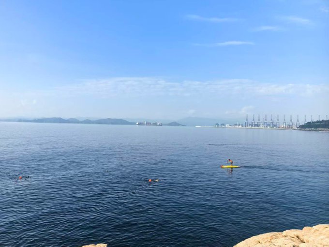A lone paddleboarder glides across calm blue waters with a distant industrial port and mountain coastline in the background, while small swimmers float nearby in the serene bay. The peaceful maritime scene provides a tranquil contrast to the post's discussion of cryptocurrency market cycles and BNB's resilience across bear and bull markets.