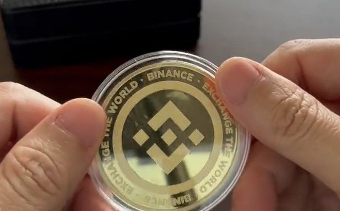 A pair of hands holds a gold Binance coin in a clear plastic capsule, with 'CHANGE THE WORLD • BINANCE • EXCHANGE THE WORLD' encircling the central logo.