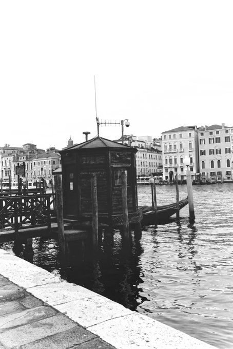 A black and white photograph of a Venetian scene featuring a wooden dock structure, a gondola moored in the water, and historic buildings along the Grand Canal in the background.
