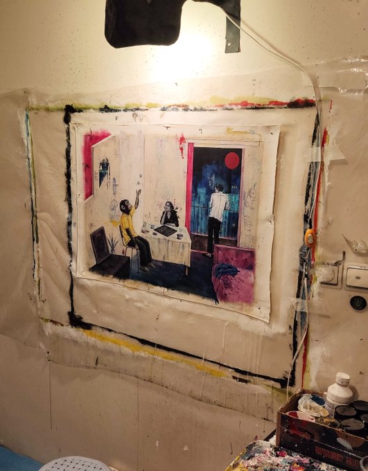 A photograph of artwork displayed on a wall, appearing to be a piece depicting people at a table with a red circular element, framed by paint splatters and tape in what looks like an artist's studio.
