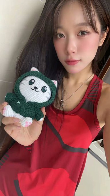 The image features a young woman wearing a red tank top, holding up a small white cat toy with a green hood. The girl appears to be posing for a photo or selfie, as indicated by her expression. The background of the image shows a bathroom setting, which might suggest that she is in a private space like a bedroom or a personal bathroom.