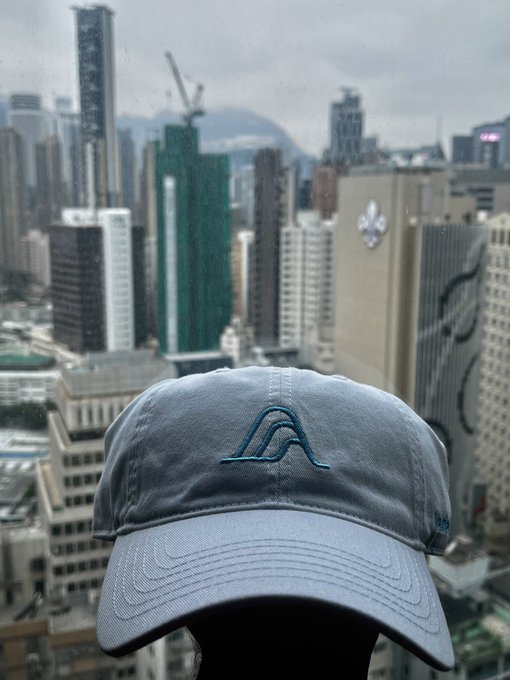A gray baseball cap with a blue embroidered logo held up against a rainy, hazy cityscape of Hong Kong's high-rise skyline.