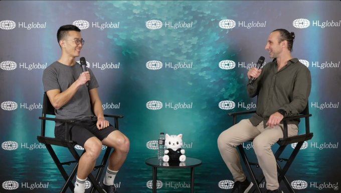 Two men hold microphones during an on-stage interview in front of a repeating “HLglobal” backdrop, with a small round table between them. The image illustrates the Hyperliquid Community Event radio session featuring Chameleon Jeff and ecosystem builders, whose 13-session recordings are available via the linked YouTube playlist.