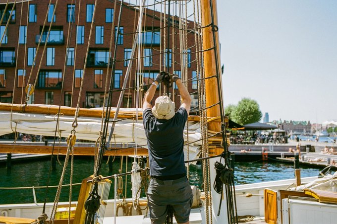 A man wearing a white cap and dark blue shirt stands on a sailboat, working on the rigging with a brick building and harbor visible in the background.