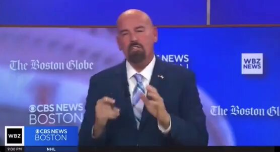 The image shows a man wearing a suit, standing at a news conference or event. He has a beard and mustache, giving him a distinguished appearance. The background of the image features a blue wall with white text that reads "The Boston Globe" and "WBCN". There is also an announcement about a cryptocurrency called Ethereum on this wall. The man appears to be speaking at the event, possibly discussing the relationship between Bitcoin and Ethereum or other cryptocurrencies.