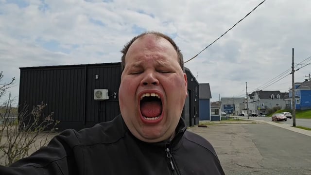 The image features a man with his mouth open, possibly yawning or panting, standing next to a building. He appears to be wearing a black jacket. The background of the photo shows an urban setting with cars parked along the street and power lines running above them. There is also a sign in the scene that reads "Calling Bitcoin in May." This suggests that the man might have been discussing or thinking about Bitcoin, possibly related to its value or market trends at the time he took this picture.