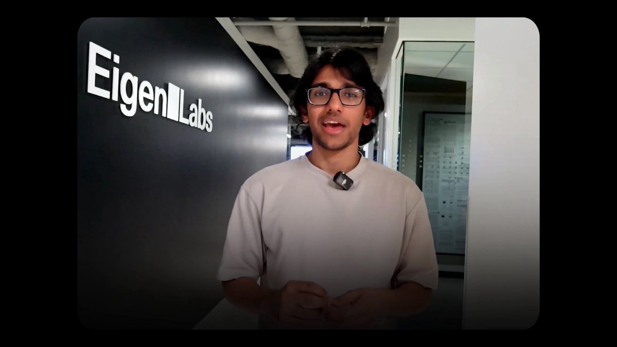 The image shows a man wearing glasses standing in front of a black wall with the word "EigenLabs" written on it. The man appears to be giving a presentation or speech, as indicated by his body language and facial expressions.