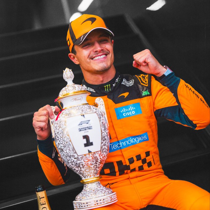 A smiling F1 driver in an orange McLaren race suit and cap celebrates with a decorative trophy marked "1" while clenching his fist in triumph, appearing ready for another victory.