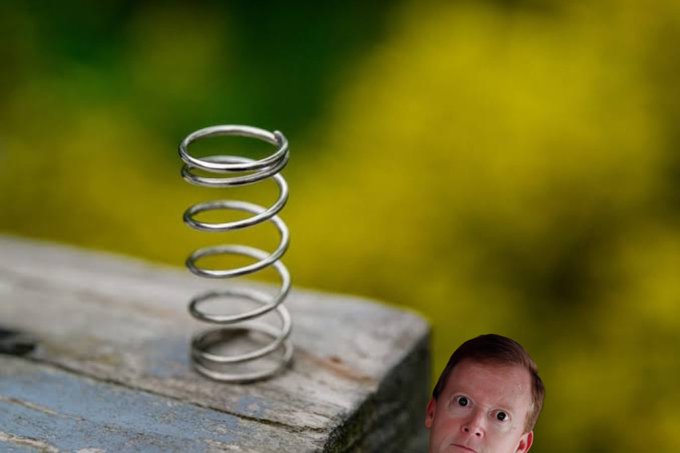 A metal coil spring sits on a wooden surface with a blurred yellow-green background, while a cutout of a surprised-looking man's face appears in the bottom right corner.
