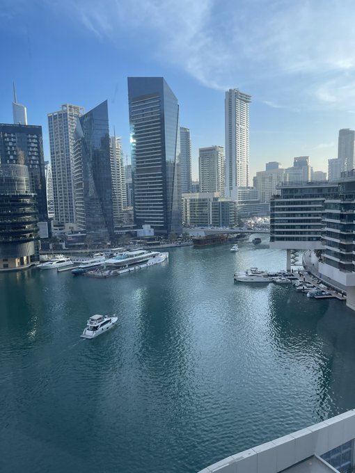 A view of a marina with boats and yachts surrounded by modern high-rise skyscrapers under a blue sky with scattered clouds.
