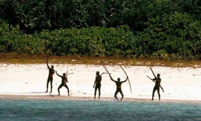 Five indigenous people stand on a white sandy beach holding bows, with dense green jungle vegetation behind them and turquoise water in the foreground.