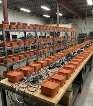 An industrial warehouse or workshop filled with rows of orange electronic equipment or mining hardware units arranged on wooden tables and metal shelving, connected by numerous cables.