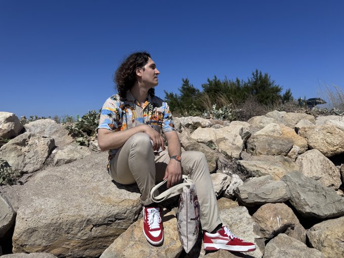 A young man with curly hair sitting on rocks outdoors, wearing a colorful printed shirt, beige pants, and red sneakers, holding a bag while looking off to the side under a clear blue sky.