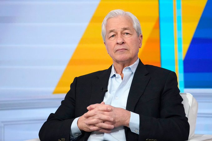 The image features an older man sitting at a desk, wearing a suit and tie. He appears to be a well-known figure or a prominent individual. The background of the image includes a colorful abstract design with yellow, blue, and orange colors. There is also text in the image that reads "jpmorgan ceo jamie dimon is pushing back hard against stablecoin rewards in the clarity act debate, warning banks will not accept”. This suggests that there might be an ongoing discussion or legal battle related to stablecoins and their acceptance by banks.