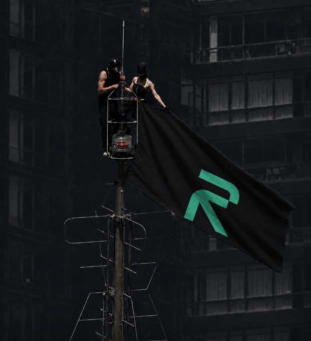 Dramatic image of two masked figures atop an antenna structure hanging a black flag with a green 'R' logo (Roblox), viewed against a dark building