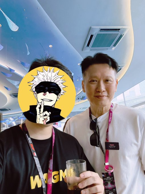Two people pose together at the Singapore Grand Prix (F1) event, wearing red VIP lanyards against a painted sky ceiling with raindrop motifs. The person on the right is Binance CEO Richard Teng, whose name badge "Richard" is visible on his white shirt, while the other person has their face covered by a Gojo Satoru anime sticker and wears a "WAGMI" shirt, holding a drink. The image ties directly to the post by confirming the casual in-person encounter with Teng at the F1 race alongside Binance team members.