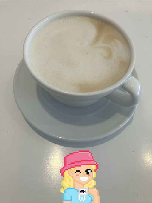 A white cup of frothy cappuccino sits on a gray saucer, with a pixel art sticker of a blonde character wearing a pink beanie labeled "GM" overlaid at the bottom.