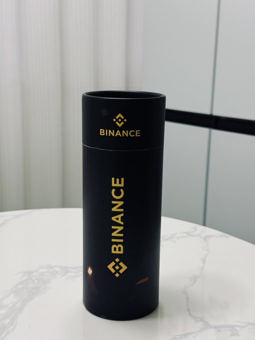 The image features a black cylindrical object with gold lettering that reads "binance" on its side, placed on top of a white table. The background appears to be a white wall or a marble countertop. There are no other objects in the scene besides the black and gold item.