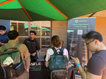 A group of attendees visits the Mantle booth under a green canopy at the StanfordSBA Science of Blockchain Conference, where a representative engages with visitors beside a banner reading "Where Real-World Finance Flows." The scene captures the in-person networking and education moment referenced in the post, showcasing Mantle's outreach to founders exploring cross-border real-world finance solutions.