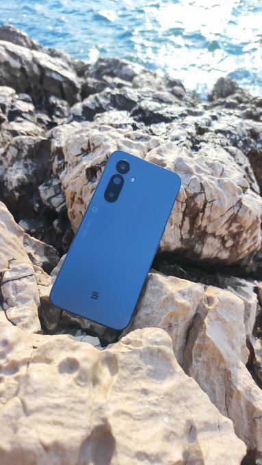 Blue smartphone (appearing to be a Xiaomi device) lying on rocky coastal terrain with ocean water visible in the background.
