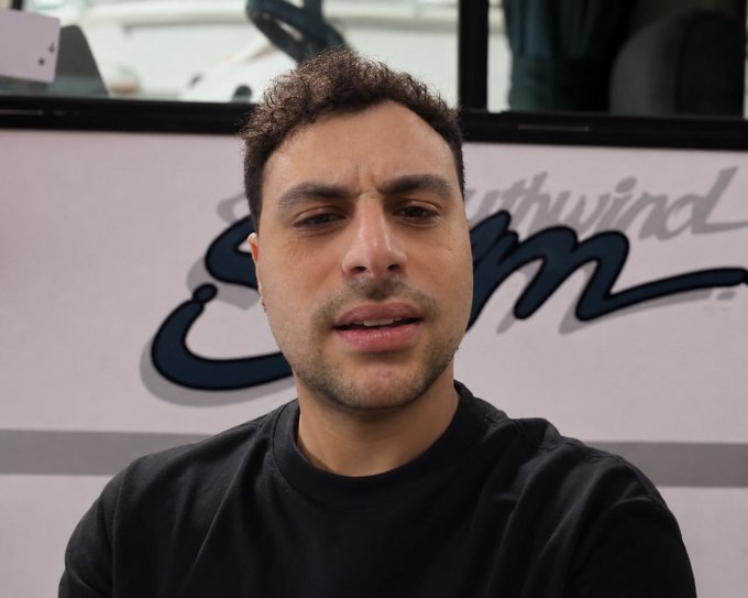 A man with curly brown hair and a black shirt speaks directly to the camera in front of a sign reading "Southwind," likely filming a video response. The image pairs with a post jokingly asking about crypto hardware wallet security, suggesting he may be reacting to or addressing concerns about Trezor and Ledger devices.
