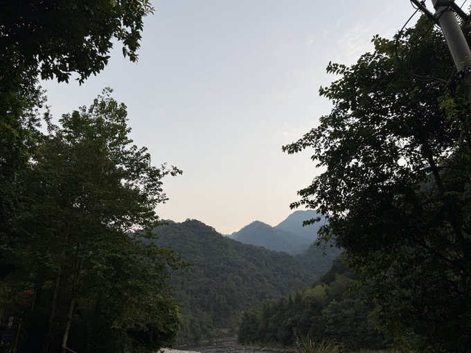 A serene mountain valley framed by lush tree silhouettes, with misty peaks rising in the distance under a soft, hazy sky—evoking the peaceful countryside retreat the poster dreams of building one day.