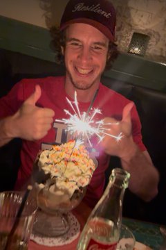 A smiling young man giving two thumbs up while celebrating with a sparkler candle on a dessert at what appears to be a restaurant.