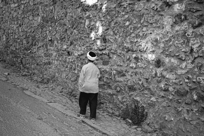 A black and white photograph of a small child wearing a beanie and oversized jacket, standing alone and looking up at a tall, textured stone wall.