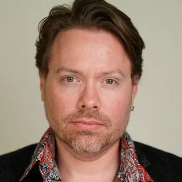 A close-up headshot portrait of a man with brown hair, blue eyes, and a small earring, wearing a dark jacket over a patterned collared shirt against a neutral background.