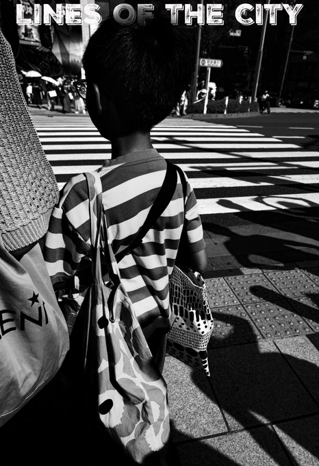 A black-and-white street photograph shows a child from behind, wearing a striped shirt and carrying bags, waiting at a crosswalk's bold painted lines, with the text "LINES OF THE CITY" overlaid at the top—directly illustrating the post's "Lines of the City" street photography theme while teasing the 0.04 ETH SuperRare NFT listing.