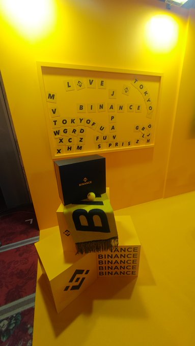 A yellow-themed Binance branded display or event space featuring a Scrabble-like tile board spelling 'BINANCE' and 'TOKYO', with a tiered podium displaying a 'B' and Binance logos.