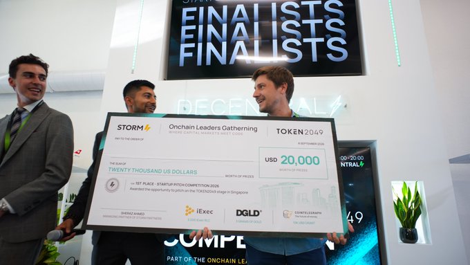 Three men smiling while holding a giant novelty check for 'Twenty Thousand US Dollars' at the Token2049 Onchain Leaders Gathering, with 'FINALISTS' displayed on a screen behind them.