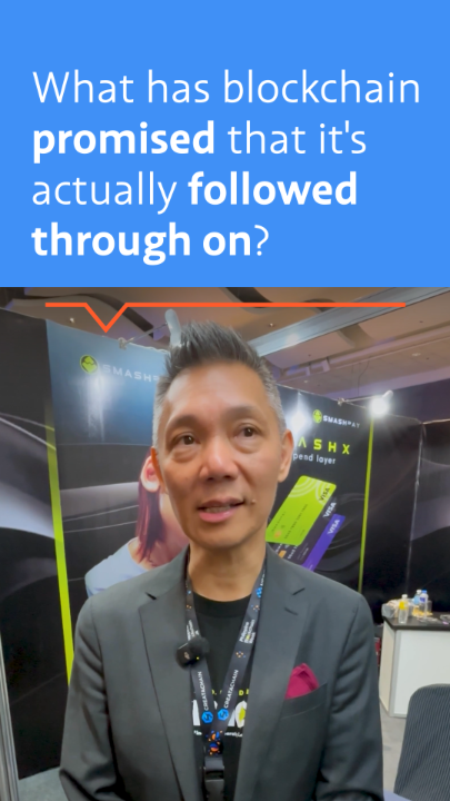 A man in a grey suit speaks on camera at what appears to be a crypto conference (with 'SMASHX' and 'GEMS' signage in the background), with a blue banner at the top reading 'What has blockchain promised that it's actually followed through on?'.