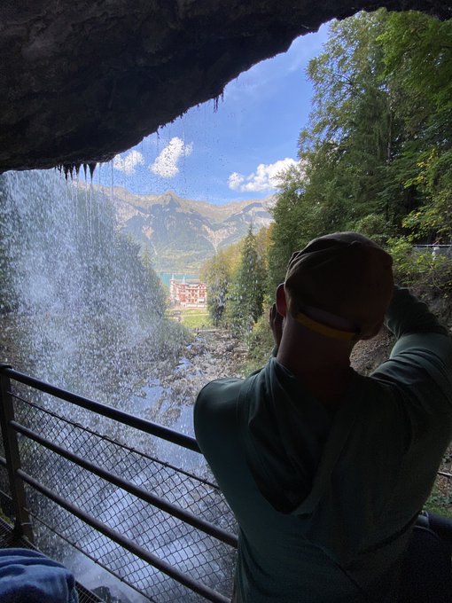 Person wearing a green hoodie and cap viewed from behind, looking out from inside a cave or rock shelter at a cascading waterfall, valley, and distant mountains.