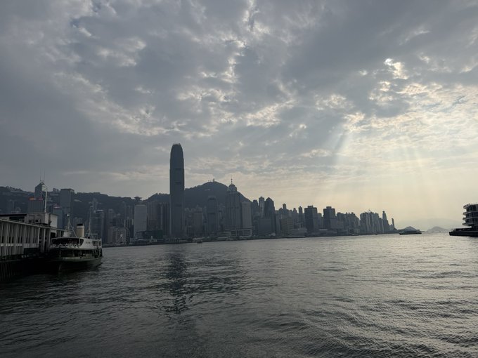 A scenic view of Hong Kong's skyline across Victoria Harbour at sunset, with sunbeams breaking through dramatic clouds and a small ferry on the water.