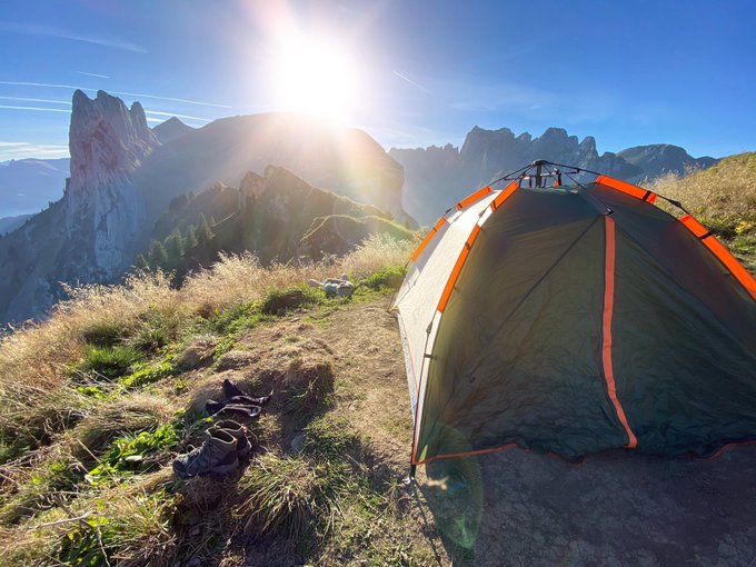 A scenic mountain landscape featuring a green and orange camping tent set up on a grassy ridge, with rugged mountain peaks and the sun rising in the distance.