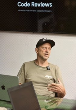 A man wearing a cap and an olive green t-shirt speaks into a microphone during a presentation, with a laptop displaying "Code Reviews: A Practical Guide for Open Source Contributors" projected on the screen behind him. The scene captures a teaching moment where open-source expert Jon Atack shares knowledge with the Bitcoin Office team and CUBO+ students, embodying the mentorship program highlighted in the post about building El Salvador's next generation of Bitcoin talent.