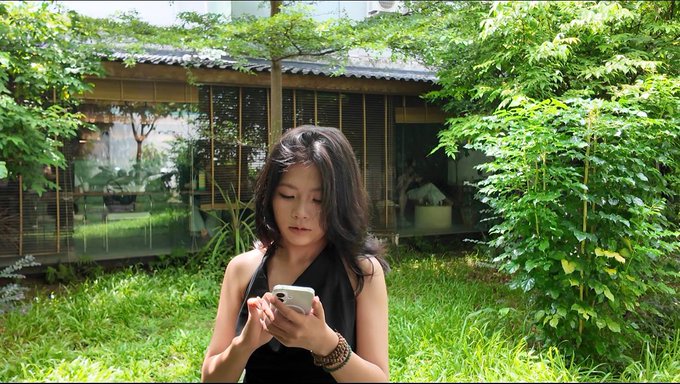 A woman with long dark hair stands in a lush green garden looking down at her smartphone, with a traditional-style building visible behind her.