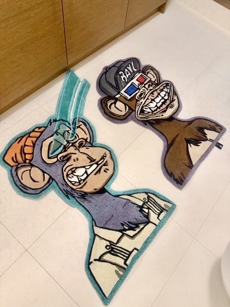Two anime-style decorative rugs are laid on a light-colored tiled floor, featuring cartoon characters with exaggerated grinning faces, one wearing a baseball cap labeled 'BAN'.