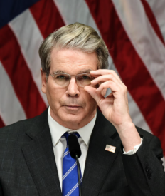 A man in a dark suit with an American flag pin and blue striped tie adjusts his glasses while speaking at a podium with a microphone, with American flags in the background.