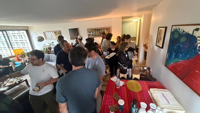 A casual gathering of people in an apartment with artwork on the walls, food, and drinks on a table covered with a red tablecloth.