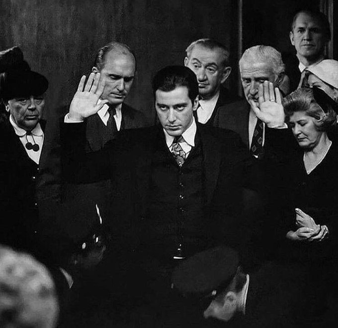 A black and white photograph depicting a swearing-in ceremony, with a central figure raising his right hand while others look on, including an older woman with white hair.