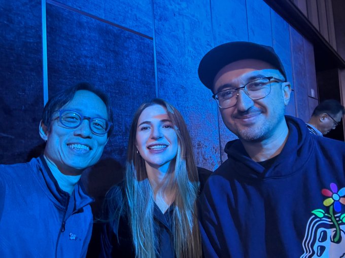 Three people pose smiling for a selfie at what appears to be an event venue bathed in blue lighting, with the man on the right wearing a hoodie featuring a colorful flower design. The image relates to the post by capturing the co-panelists mentioned, likely including moderator Jacq Melinek alongside panelists Ysiau and Mfigge, during their discussion at the KBW (Korea Blockchain Week) event about the scarcity of human creativity in an age of abundant intelligence.