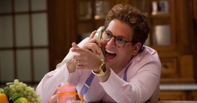The image features an actor dressed in a pink shirt, holding a phone up to his ear with both hands while laughing. The background includes a dining table with various items such as grapes, oranges, and a cup. There are also two clocks visible in the scene - one on top of the dining table and another smaller clock near the bottom right corner.