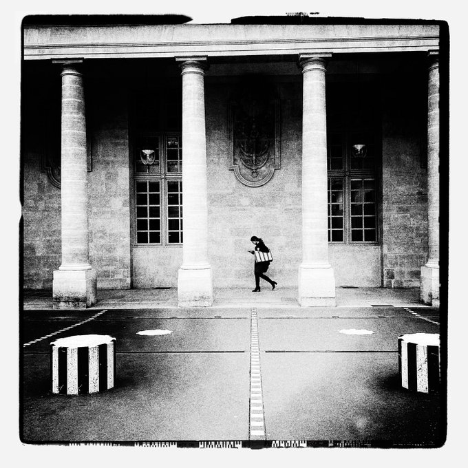 The image shows a person walking across a classical building's facade, which has columns and windows. The person appears to be crossing the street or moving through an intersection. In the foreground of the photo, there are two striped bags that match the geometric pattern of the building's architecture. The image also contains text elements such as "GM Excellent Sunday Assorti au sac de a black-and-white piece where a solitary figure crosses a classical façade, with the striped bag echoing the geometry of the scene."