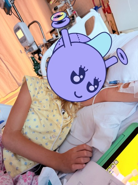 A young patient in a yellow floral hospital gown lies in a hospital bed with medical equipment and an IV visible; their face is obscured by a large purple cartoon bee sticker.
