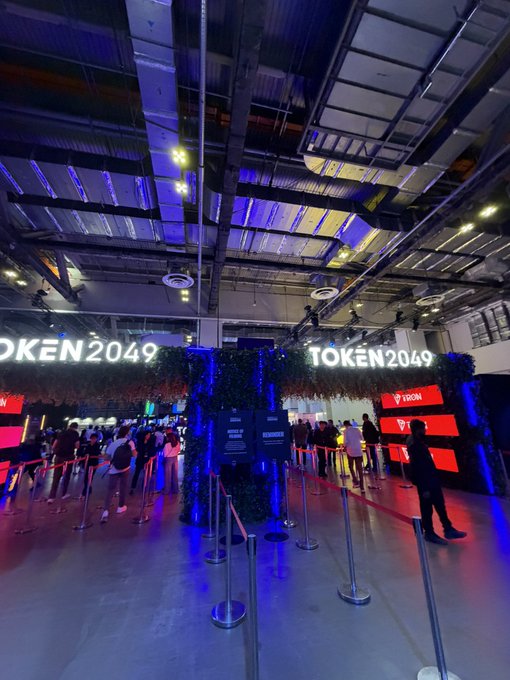 The entrance area of TOKEN2049 conference, featuring the event's name illuminated on large signs overhead, with attendees queuing behind stanchions and red ropes beneath a high industrial ceiling lit with blue and purple ambient lighting.